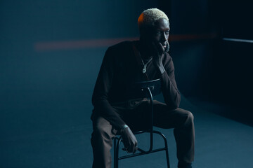 dark-skinned handsome guy with white hair and blue eyes sitting on a chair in a dark studio with a serious facial expression in brown clothes and silver accessories, he looks away - 412130198