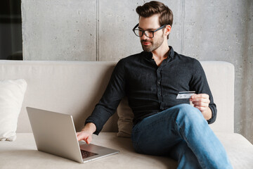 Focused unshaven man using laptop and credit card while sitting on couch
