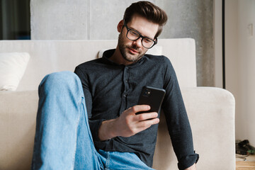 Serious unshaven man using smartphone while sitting on floor