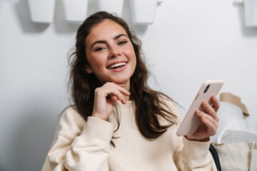 Joyful girl smiling and using mobile phone while sitting indoors