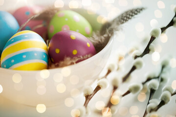 holidays and object concept - colored easter eggs with quail feathers in bowl and pussy willow branches over bokeh lighs