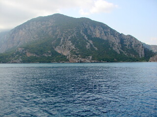 Landscape of the calm azure surface of the sea bay surrounded by mountain forests under a cloudy morning sky.