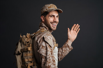 Smiling masculine military man waving hand while posing with backpack