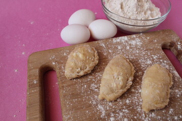 Raw cooking dumplings lie on a board beside egg flour on a pink background view from above