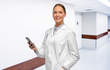 medicine, profession and healthcare concept - happy smiling female doctor with stethoscope using smartphone over hospital corridor background