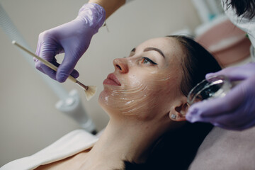 Beautician applying with brush contact gel on face of woman for hardware procedures in beauty spa