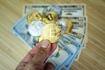 Bitcoin (btc, xbt) coin in golden shiny metallic which is holding on the people hand with blurred stack of many coins and US dollar as background on the wooden table.