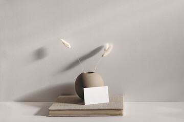 Modern stationery still life scene. Beige spheric vase with dry lagurus grass. Table background in sunlight. Blank business card mockups lean on old book. Champagne wall, long shadows.