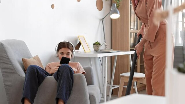 Wide Shot Of Teenage Caucasian Girl Wearing Headphones Lying In Armchair With Phone In Hands While Her Mom Vacuuming The Floor In Apartment