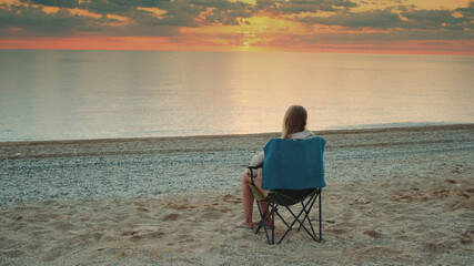 Fototapeta premium Woman sitting on camping chair and drinking coffee. Relaxing and enjoying nature. Admiring sunrise on the sea.
