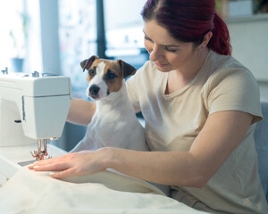 Caucasian woman sews while sitting in the kitchen. Dog Jack Russell Terrier sits on the lap of the...