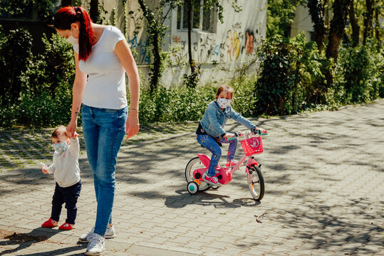 Young Mother Wearing A Mask Walking One Daughter On Foot And Big Sister On A Bicycle Outdoors During The Pandemic Year Of 2021