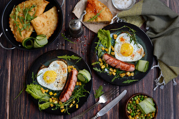 Heart shaped scrambled eggs and sausages with vegetables on dark plates on a dark wooden background. Top view