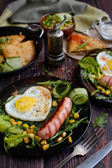 Delicious and healthy breakfast. Festive breakfast for Valentine's Day, Mother's Day. Heart shaped fried eggs and fried sausages with herbs and vegetables on a brown wooden background.