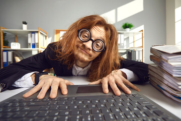 Man sitting at office desk and typing on keyboard. Funny nerd with long ginger hair searching for information on the Internet. Crazy looking computer geek in round thick lens glasses working on laptop - obrazy, fototapety, plakaty