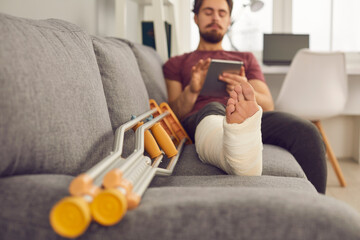 Young man with broken leg in plaster cast sitting on sofa and using tablet computer. Close up of...
