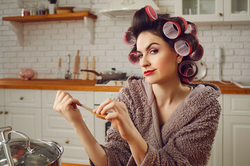 Beautiful woman wearing makeup, funny hair curlers and housecoat sitting at kitchen table at home,...