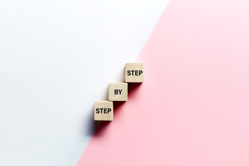 The words step by step on wooden cubes against pink and white background. Progress or growth in...