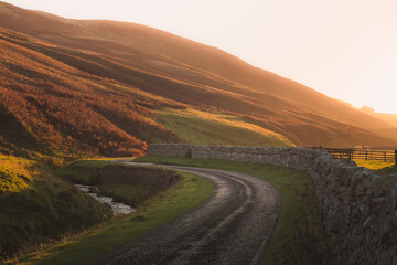 Golden sunset or sunrise light on an empty country road alongside an old stone wall on the Glencorse walk in the Pentland Hills Regional Park countryside in Edinburgh, Midlothian, Scotland. © Stephen