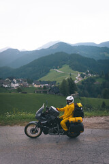 A girl in a yellow raincoat, shoe covers and a motorbike helmet. Motorcyclism and travel. Vertical photo. Top of the Mountains. A day with clouds. Difficult test and extreme vacation. Biker outfit.