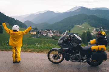 A happy girl in a yellow raincoat, shoe covers and a motorbike helmet. Motorcyclism and travel. Top of the Mountains. A day with clouds. Difficult test and extreme vacation. Hands up