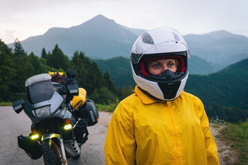 Fototapeta na wymiar Close-up view. Woman in a yellow raincoat, a white helmet. Motorcyclism and travel. Sightseeing tour. Mountains and hills. A gray day with thunderclouds. Difficult test and biker's outfit.