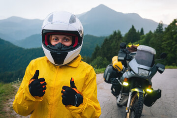 Close-up view. Happy woman in a yellow raincoat, a white helmet. Motorcyclism and travel. Fingers up, take like. Mountains and hills. A gray day with thunderclouds. Difficult test and biker's outfit.