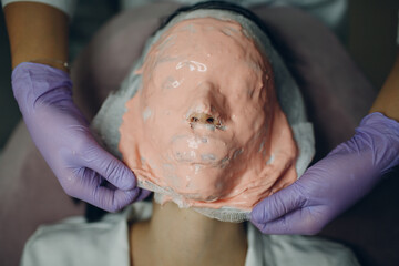 Alginate face mask removal process. A female hands of a beautician holding spatula. Woman lying on a couch in office of cosmetologist. Facial skincare in the beauty spa salon