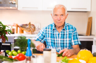 elderly man cuts vegetables for salad at the table in the kitchen