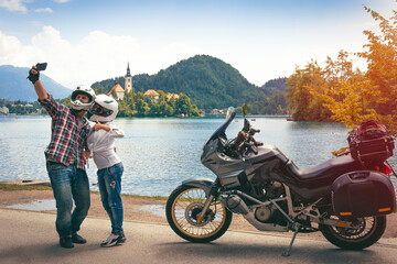 Fototapeta na wymiar Couple in love travel together takes a selfie on a smartphone. Stands with motorcycle with bags. Tourism and vacation. Sunny day. Bled lake, island castle and mountains in background, Slovenia Europe