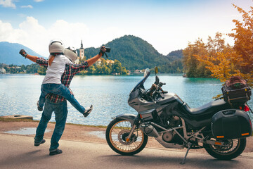 Fototapeta na wymiar Happy fun biker couple in love travel together. Stands by a motorcycle with bags. Tourism and vacation. Sunny summer day. Bled lake, island, castle and mountains in background, Slovenia, Europe
