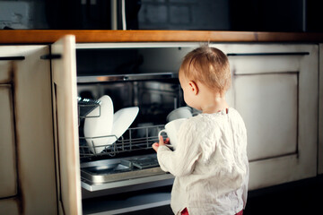 child toddler unloads the dishwasher on his own in a Scandinavian-style kitchen, child's mother's helper, housework with the child, household chores, household