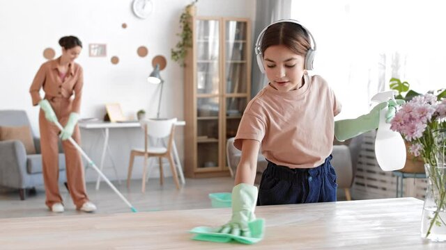 Lockdown of lovely Caucasian girl wearing earphones and gloves wiping desk using rag and sprayer while her mother mopping floor on background