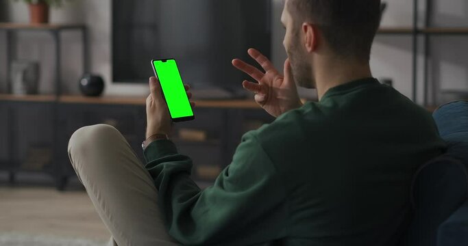 man is chatting online by video chat in mobile phone, sitting at home on couch in living room, green screen of cellphone, chroma key technology