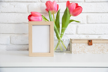 Wood frame mockup with soft pink tulip flowers in vase near painted brick wall. Empty frame mock up for presentation design. Template framing for modern art. Wooden box. Modern white chest of drawers.