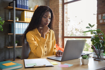 Photo of thoughtful dark skin woman browse internet laptop on table think business plan wear yellow...