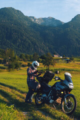 Man motorcyclist Looking for something in the trunk. Alpine mountains on background. Biker lifestyle, world traveler. Summer sunny sunset day. Green hills. hermetic packaging bags. Vertical photo.