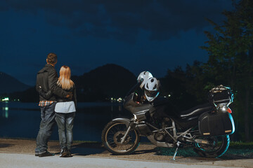 Fototapeta na wymiar Motorcyclists couple in love travel together. Stands by a motorcycle with bags. Tourism and vacation. Summer night. Bled lake, island and mountains in background, back view. Slovenia, Europe