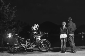 Fototapeta na wymiar Motorcyclists couple in love travel together. Stands by a motorcycle with bags. Tourism and vacation. Summer night. Bled lake, Black and white, back view. Slovenia, Europe