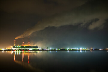 Brightly illuminated coal power plant high pipes with black smoke moving upwards polluting atmosphere at night with reflections of it in lake water.