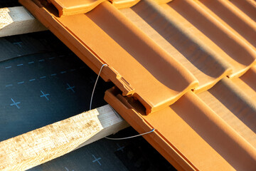 Closeup of metal montage anchor for installation of yellow ceramic roofing tiles mounted on wooden boards covering residential building roof under construction.