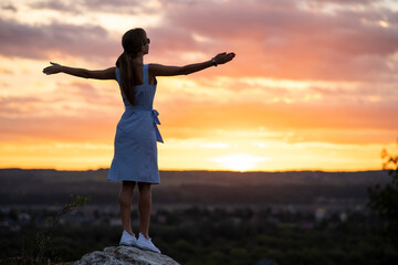 Fototapeta na wymiar A young woman in summer dress standing outdoors with outstretched arms enjoying view of bright yellow sunset.