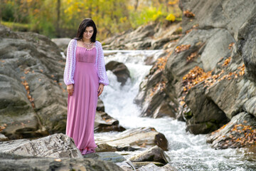 Young pretty woman in long fashionable dress standing near small mountain river with fast moving water.