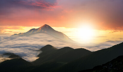 Sunset landscape with high peaks and foggy valley with thick white clouds under vibrant colorful evening sky in rocky mountains.