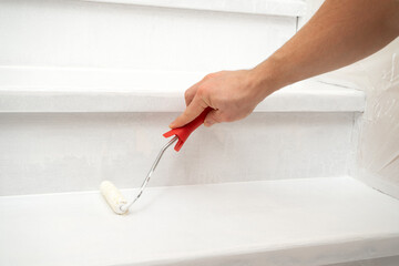 male hand paints a wooden staircase white with paint and a roller.