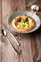 Hummus in a grey bowl seasoned with sunflower and pumpkin seeds, sesame, olives and olive oil. Close-up on a table with cutlery, seeds and garlic by side. Isolated dish on wooden background.