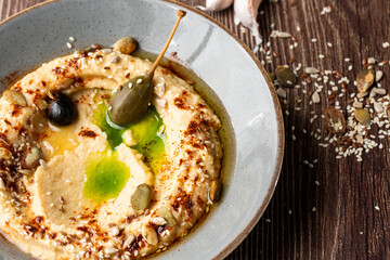 Hummus in a grey bowl seasoned with sunflower and pumpkin seeds, sesame, olives and olive oil. Macro close-up on a table with seeds and garlic by side. Isolated dish on wooden background.