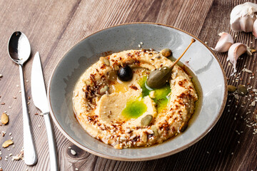 Hummus in a grey bowl seasoned with sunflower and pumpkin seeds, sesame, olives and olive oil. Close-up on a table with cutlery, seeds and garlic by side. Isolated dish on wooden background.
