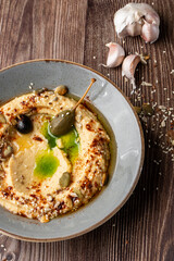 Hummus in a grey bowl seasoned with sunflower and pumpkin seeds, sesame, olives and olive oil. Macro close-up on a table with seeds and garlic by side. Isolated dish on wooden background.