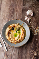 Hummus in a grey bowl seasoned with sunflower and pumpkin seeds, sesame, olives and olive oil. Close-up on a table with cutlery and seeds by side. Top view isolated on wooden background. Copy space.